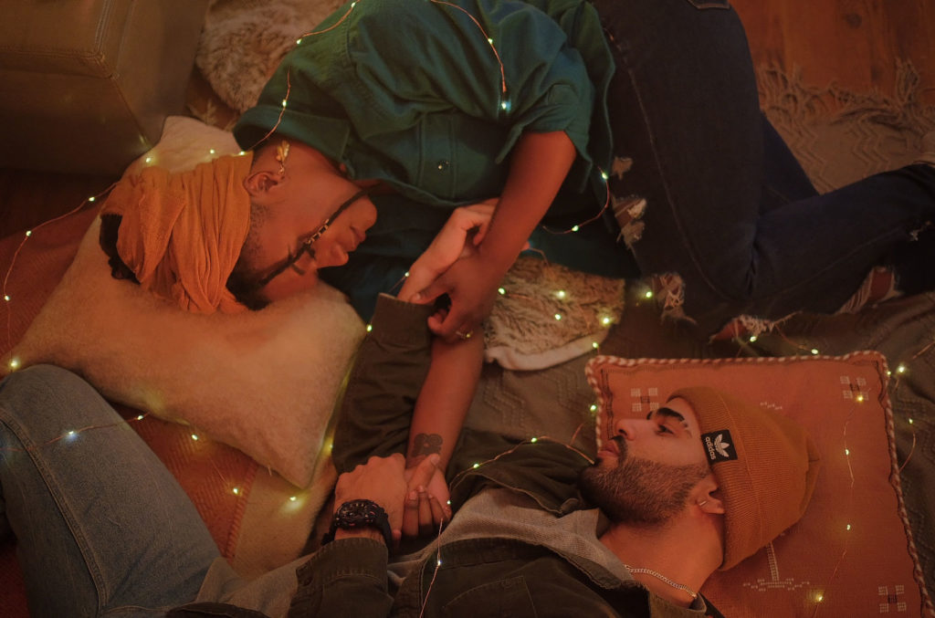 A couple holds hands on the floor, on pillows, with a string of lights around them