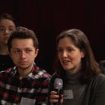 A presenter speaking into a microphone while some nearby observers look on