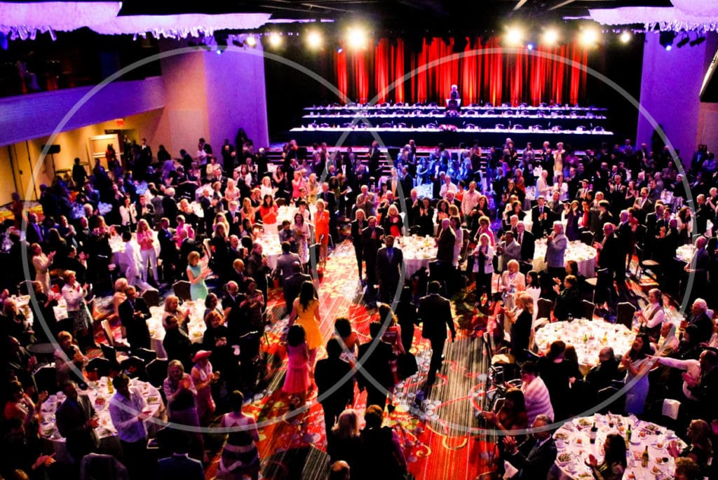 Wide shot of Drama League Gala, hundreds of patrons in foreground with red curtain and stage in background.