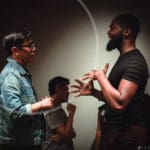 Two artists chatting and using their hands to communicate in a dark room with another artist in the background